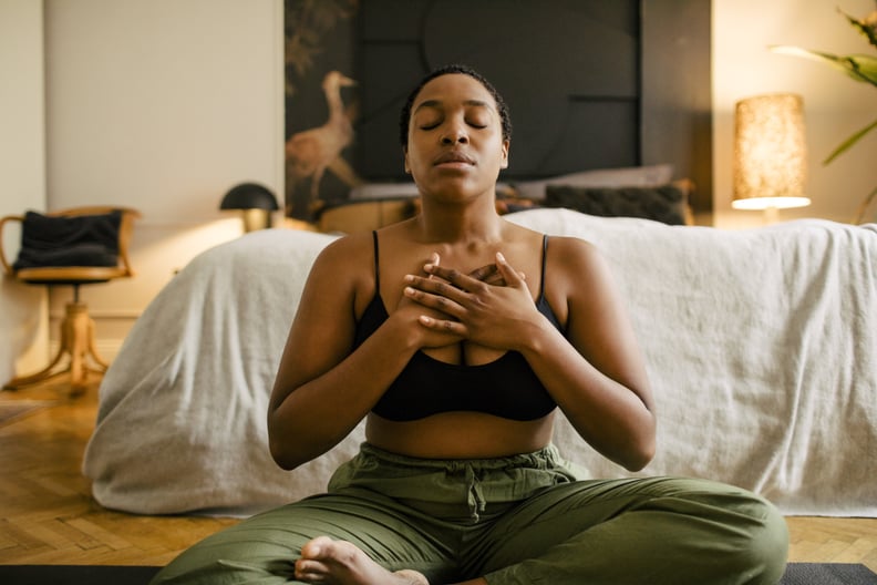 A Black woman sits cross-legged in a bedroom with her hands over her chest in a meditating position.